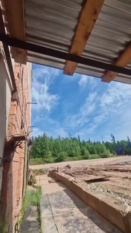The video pans from left to right, showing a view from under a partially constructed roof. The roof is made of corrugated metal supported by wooden planks and metal beams. To the left, a brick wall is visible. The foreground is a dirt and gravel area with concrete barriers. In the background, a dense green forest with some taller trees is seen under a bright blue sky with wispy white clouds. A dirt road winds through the landscape, and in the distance, there appears to be a construction site with some structures and possibly a vehicle. The overall impression is of an outdoor, rural, or industrial setting under sunny weather.