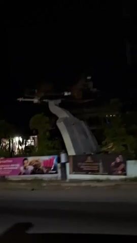 The video captures a nighttime outdoor scene, likely from a moving vehicle. In the midground, a large monument features a dark-colored fighter jet airplane mounted on a prominent white, curved support structure. Below the monument and along the side of the road, multiple illuminated billboards and advertisements are visible, displaying images of people and text in various colors like pink, blue, and yellow. Trees and bushes are present around the base of the monument and along the road. Distant buildings with some lights are visible in the background against the dark sky. The camera pans slowly from left to right, following the monument as if passing by it.