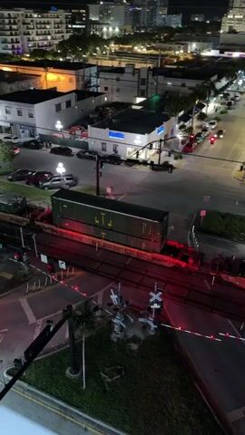 A train is stopped at a railroad crossing at night. The train is long and green, with multiple cars. The crossing lights are flashing red and white. There are several cars and trucks on the road near the crossing. There are also a few people standing near the train. The background shows a city skyline with buildings and streetlights.