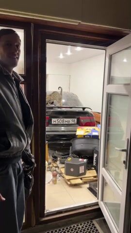 A man stands in a doorway, holding a black plastic bag, and points into an indoor space, revealing a black Opel Calibra. The car is covered in dust, and sitting on the back deck are two cats, one tabby and one black. The garage is cluttered with boxes and objects, illuminated by ceiling lights. A woman's arm is visible, also pointing towards the cats on the car.
