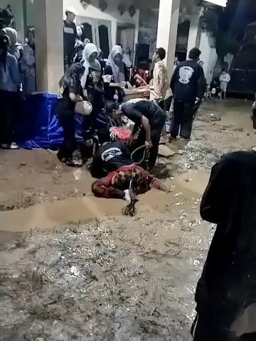 At night, a group of people are gathered on muddy ground in Jenggrik, Indonesia. In the foreground, a person dressed in traditional patterned clothing lies prone in a puddle of mud. Standing over them is another individual in a black t-shirt, holding a whip or riding crop. Surrounding them are several other people, mostly young adults, observing the scene. Some women wear headscarves, and many individuals are dressed in dark t-shirts, some with white graphics or text on the back that are not clearly legible. A small wooden table or stool is present near the central figures, along with a blue tarp or fabric on the left. The setting appears to be outdoors, likely in a rural or village area, with buildings visible in the background. The muddy conditions suggest a wet environment or recent rain. The scene portrays what appears to be a cultural performance or ritual, possibly a Jaranan trance dance, given the attire, actions, and local context.
