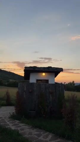 A small, white building with a dark, tiled roof is centered in the frame, illuminated by a single yellow light above its doorway. The building is surrounded by a rough, stone-like enclosure. Several small, evergreen trees are planted around the enclosure. A path made of uneven, light-colored stones leads up to the structure. The background shows a gently sloping hill covered in trees on the left, and a field on the right. The sky above is a gradient of pale blue and orange, indicating sunset or sunrise. There are no people visible in the video. The video is a still image with no discernible action.