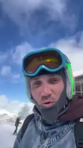 A man wearing a blue and green ski helmet, orange goggles, and a gray neck gaiter is holding a smartphone camera in front of him, recording a selfie video. He is wearing a denim jacket over a brown hoodie and has a black backpack on. In the background, there is a snowy scene with trees, and other people skiing. At one point, he smiles, sticks his tongue out and waves his gloved hand at the camera.