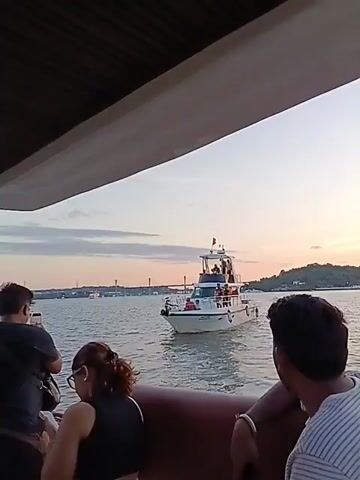 The video shows a white motorboat moving on the water, filmed from another boat, likely during sunset. The motorboat has people on board, some standing on the upper deck. In the background, a bridge is visible, along with some land. The water is slightly choppy. The camera is held by someone on another boat, as other people can be seen on the edges of the frame. One person is taking a picture with their phone.