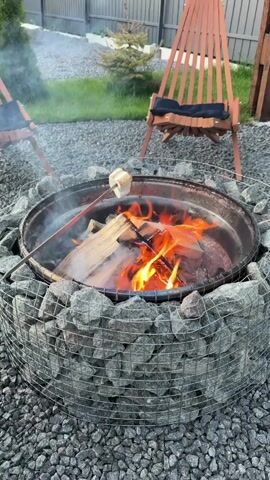 A marshmallow is being toasted over a campfire in a gabion fire pit. Two chairs are visible in the background. The fire pit is surrounded by gravel.