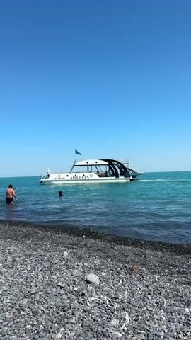 A man stands in the shallows of a turquoise lake, facing away from the camera, with a boat named "AISULTAN" visible in the background. Another person is swimming in the water between the man and the boat. The boat appears to be a tour boat with passengers visible on its upper deck, and a slide extending from the back. The shore is covered in dark grey and black pebbles. The sky is clear and blue.