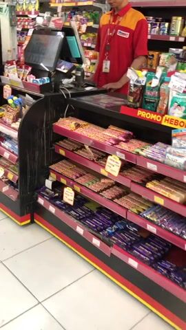 The video shows a cashier in a red and yellow uniform standing behind a checkout counter in a convenience store. The counter has a touchscreen monitor and a card reader. Shelves stocked with candy bars are visible in the foreground, with a "PROMO" sign displayed on the right side of the counter. The store appears to be well-lit and organized, with a variety of products on display.