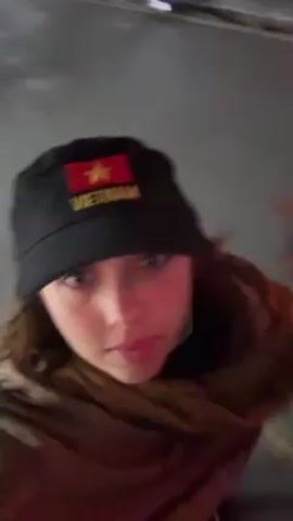 A person is wearing a black bucket hat with the flag of Vietnam and the word "VIETNAM" embroidered on it in yellow thread. They are wearing a dark jacket and a brown scarf. The person is holding the brim of their hat with one hand. The background appears to be a dimly lit gray wall. The camera angle is from a slightly low perspective looking up at the person.