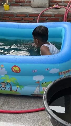 A young child, likely a boy, is sitting in an inflatable, blue, rectangular kiddie pool filled with water. He is wearing a white tank top. The pool is decorated with cartoonish images of a monkey, sun, sailboat, alligator, and butterflies. A red hose is nearby on the ground, and a black bucket filled with water sits to the right of the pool. In the background, there is a brick wall and a potted plant. The child picks up a small red toy and holds it above his head before placing both hands behind his head, resting them on the edge of the pool. The setting appears to be an outdoor residential area.