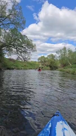 The video shows a first-person perspective from a kayak moving down a narrow river. Another kayaker in an orange kayak is visible ahead, paddling in the same direction. The river is bordered by grassy banks with trees on either side. The sky is blue with scattered white clouds. The water is calm with slight ripples. The front of the camera's blue and white kayak is visible in the foreground.