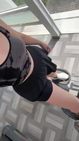 A woman is exercising on an elliptical machine in a bright, sunlit room. She is wearing a black and grey tie-dye sports bra and black shorts with white sneakers. The elliptical machine is silver with black accents. The floor is tiled in a grey herringbone pattern. The room has large windows.