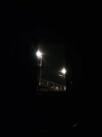 The video displays a dark outdoor environment at night, visible through a rectangular opening. The scene is illuminated by two streetlights positioned on a pole. One light is higher and to the left, while the other is lower and to the right, casting a yellow glow. Below the lights, a railing and a dark surface, possibly a pathway or stairs, are dimly visible. The overall impression is that of a gloomy, deserted area.