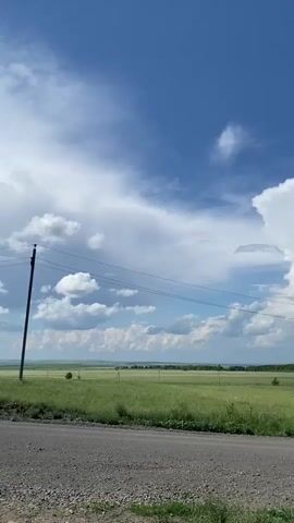 A vertical video shows a vast, flat landscape under a bright blue sky with scattered white clouds. In the foreground, a rough, gray gravel road runs horizontally across the bottom of the frame. To the left of the road, lush green grass grows. A tall, thin wooden utility pole with wires stretching horizontally across the scene stands to the left. In the middle ground, a wide expanse of green grassland stretches to the horizon, dotted with a few scattered trees and what appear to be more utility poles. The background consists of rolling, distant hills covered in greenery. A large, dramatic cumulus cloud formation, resembling a towering mushroom, dominates the sky in the center and right of the frame, its white mass contrasting with the deep blue sky. The light suggests it is daytime, possibly mid-afternoon. No people or animals are visible. The overall impression is one of rural, open space under a dynamic sky.
