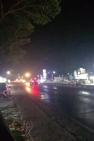 The video captures a night scene on a wet road with streetlights illuminating the area. Cars and motorcycles are moving along the road, their headlights and taillights creating streaks of light. A gas station is visible on the right side of the road, with bright lights indicating ongoing operations. Trees are present on the left side of the frame, their dark silhouettes contrasting with the lit surroundings. The overall atmosphere is one of a busy, albeit dark, urban environment, possibly after rain due to the wet appearance of the road surface.
