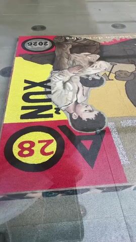 This media file is a short video capturing a view of a vibrant street art installation on the ground. The artwork features stylized illustrations of people, rendered in a semi-realistic, sepia-toned style. There are at least three distinct figures visible, depicted in various poses, suggesting a narrative or thematic composition.

The artwork is laid out on a paved surface, likely a public walkway or plaza, given the modern architectural elements in the background. The installation is rectangular, with bold, colorful blocks of red, yellow, and black serving as backdrops and borders for the illustrated figures.

Visible text elements are incorporated into the artwork. One section prominently displays the number "28" within a red circle. Another section features the text "XUN" in large, bold, yellow letters. Above the illustrations, a banner-like element has text in a cursive font, with part of it appearing to read "Letra do mestre" (which translates to "Master's Letter" in Galician, fitting the Pontevedra, Spain location context). A year, "2025", is also visible.

The artwork seems to be laid out flat on the ground, and the camera angle is from a slightly elevated perspective, looking down and across the installation. The lighting suggests it's daytime, with even illumination across the artwork. There are reflections on the surface of the artwork, indicating a smooth, possibly lacquered or glossy finish, or perhaps a layer of protection over the art. The overall impression is a dynamic and visually engaging piece of public art.