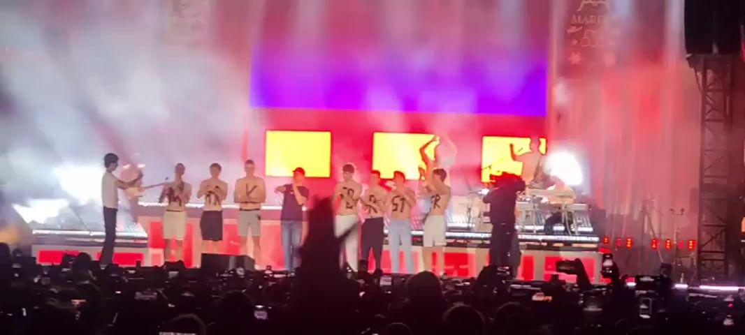 A stage is illuminated by bright lights and filled with smoke. A group of young men, mostly shirtless and wearing shorts, stand across the stage, each with a letter painted on their chests, spelling out a word. In the foreground, a crowd of people is visible, many holding up their phones to record the event. On the right side of the stage, a large screen displays a close-up of an excited audience. The overall atmosphere appears to be a concert or a live performance.