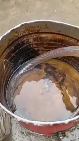 The scene unfolds outdoors on a concrete surface, revealing a red and grey metal barrel filled with murky water. A semi-transparent, ribbed hose descends into the barrel. The ground surrounding the barrel is littered with red and green plastic bottle caps. Further along the hose, a small gas-powered water pump rests on the ground, its engine red and silver. Beyond the pump, a stack of blue plastic containers with white lettering rises against a wall. Several people are present in the scene. At the top of a set of steps, an older man in a white shalwar kameez holds a white package. A man with a beard, wearing a black shalwar kameez, stands nearby. A young boy in a beige shalwar kameez leans against a parked motorcycle. The scene presents a tableau of activity, possibly involving the transfer or storage of liquid.