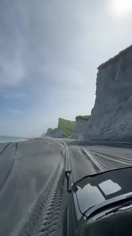 This video shows a first-person perspective of a vehicle driving on a black sand beach. The vehicle is a dark-colored SUV, and its hood and part of the windshield are visible. The beach has tire tracks from previous vehicles. To the right of the beach, there are dramatic, sheer grey cliffs with green grass on top. These cliffs recede into the distance, creating a scenic coastline. The ocean is visible to the left of the beach, with gentle waves. The sky is blue with scattered white clouds, and the sun is bright, casting a slight glare in the upper right corner of the frame. The overall scene depicts an off-road adventure along a rugged and beautiful coastal landscape.