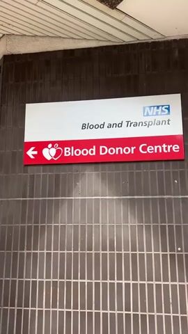 A video shows the exterior of the NHS Blood and Transplant Blood Donor Centre. A brown tiled wall has a sign with a red background, a heart with a drop of blood, and a white arrow pointing left. The sign reads, "Blood Donor Centre." To the left of the sign, there are windows with signs featuring a heart with a drop of blood and the words "Give Blood." A sidewalk is in front of the windows. The building has a white ceiling with brown trim. The camera pans to the left, revealing a long sidewalk and the side of the building. A vehicle is visible in the distance.