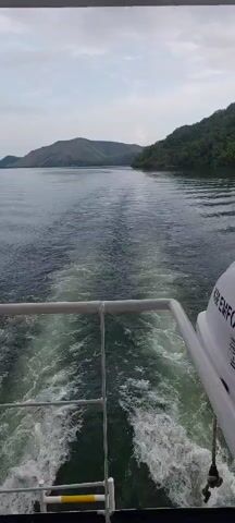 The video shows a view from the back of a moving boat. Water churns behind the vessel, creating a white wake that stretches into the distance. On the right side of the frame, part of a white life raft is visible, with text printed on it. The water is calm and appears to be a large body of water like a lake or sea. On either side of the water, lush green hills and mountains covered in trees rise up from the shore. The sky is overcast with some clouds. As the video progresses, the view pans slightly to the right, revealing more of the boat's deck and some passengers. One woman in the foreground is wearing a pink hijab, a colorful long-sleeved shirt with a tropical print, and blue jeans. She is carrying a black quilted handbag. Another person wearing a pink hijab is partially visible behind her. An orange bag is also visible on the deck. The boat appears to be traveling along a scenic route.