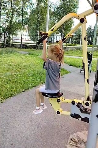 A young girl with blonde hair in a ponytail is sitting on a yellow exercise machine on an outdoor playground. She is wearing a gray t-shirt and white shorts. Her hands are on the handles of the machine, and she is pulling them down. The machine is yellow and gray, with some rust and graffiti on it. In the background, there are trees, grass, and buildings. A stroller is parked behind the exercise machine.
