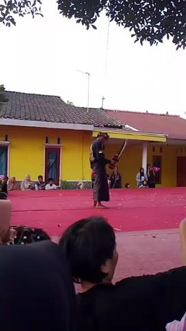 A group of people are gathered on a red carpeted stage area outside a yellow building, watching a performance. In the center of the stage, a person dressed in traditional dark clothing, possibly a dancer or performer, is in motion. They appear to be holding or manipulating a prop that is coiled and dark in color. In the background, several other individuals are seated or standing, observing the performance. The environment appears to be outdoors, with trees visible overhead and a house or building in the background. The audience in the foreground includes people of various ages, some of whom are recording the event with their phones. The overall atmosphere suggests a cultural or community event.