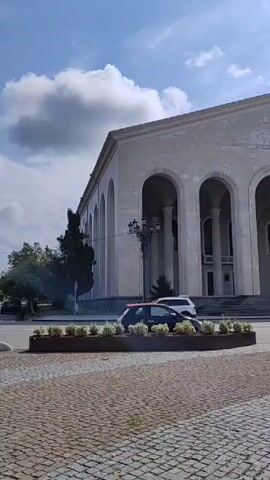 A static shot of a large, light-colored building with arched entrances and columns is captured. In front of the building, a paved area with cobblestones leads to a planter box filled with green foliage. Cars are seen driving by on the street in front of the building. The sky above is partly cloudy with a blue sky visible. The overall scene suggests an outdoor view of a public building or monument.