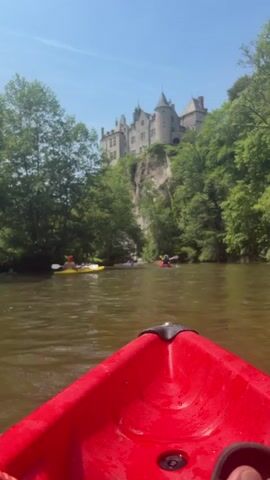 The video shows a river view from a red kayak, with a large stone castle perched atop a cliff in the background. Several people are paddling kayaks in the river, including one in a yellow kayak and another in a red one. The trees line the riverbanks and surround the cliff, while the sky is clear and blue. The camera moves slightly, adjusting the angle to capture the castle and the kayakers. The river appears calm with a muddy color.
