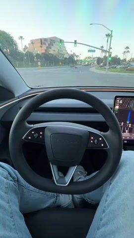 The video shows a first-person view from the driver's seat of a Tesla. The steering wheel is visible in the foreground, with the driver's legs and blue jeans underneath. The dashboard features a large central touchscreen displaying a navigation map. Outside the car, palm trees and a building are visible, along with traffic lights and a road. The sky is a light blue with some colorful clouds. The driver's hand appears in frame at 00:03, waving. The car appears to be in motion on a road.