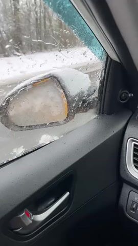 A video filmed inside a car driving on a snowy road shows the driver's perspective, with snow and ice accumulated on the car's exterior and windshield. The car's windshield wipers are active, clearing the snow from the view. The side mirror is covered in ice. The car interior shows the steering wheel, dashboard, and door panel. The road ahead is visible through the windshield, with trees lining the sides of the road and a light snowfall occurring.