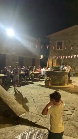 The video shows a nighttime scene in a village square. In the foreground, a young boy in a white t-shirt and blue shorts stands with his back to the camera, looking towards the right. The square is paved with cobblestones and illuminated by overhead string lights and a street lamp. In the background, a stone building with a well in front of it is visible. A group of people are gathered around tables set up in the square, some eating and socializing. Red and white pennant banners are strung across the buildings, suggesting a festive event. Several adults and children are present, and the atmosphere appears lively.