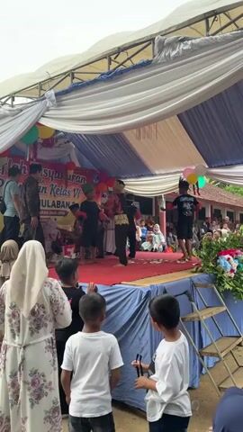 A group of children are performing on a stage covered with a red carpet and draped with white and blue fabric. The stage has a banner that reads "Pusat Pelayanan Al-Qur'an Kelas VI Mulaban 2023". Some children are wearing costumes with headdresses and traditional clothing. They appear to be dancing or acting. An audience of adults and children are watching from below. Balloons are hanging from the tent above the stage.