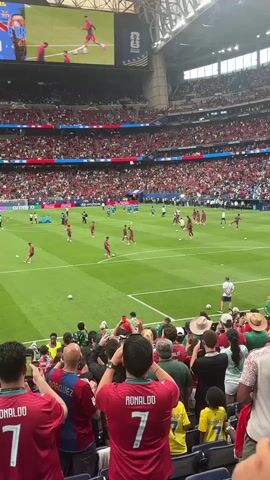 The video shows a crowd of people at a soccer game. The stadium is large with many levels of seating. The field is green and the players are warming up, some in red uniforms and some in blue. The fans are wearing various colors, with many wearing red. Some people in the foreground are holding up their phones to record the event. The atmosphere appears energetic and exciting.