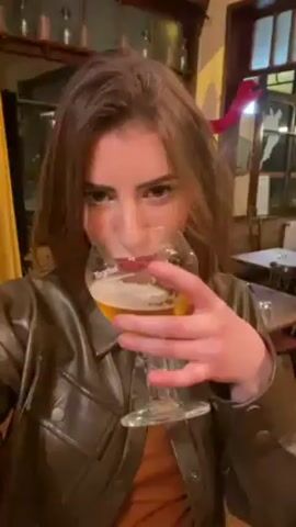 The video shows a woman with brown hair, wearing a brown leather jacket and brown shirt. She's indoors in what appears to be a restaurant or bar. She holds a glass of beer, takes a sip, and then puckers her red lips toward the camera. The camera zooms in very close to her face. The background shows tables, chairs, a window, and shelves holding items that could be glasses or trophies. The lighting is warm.