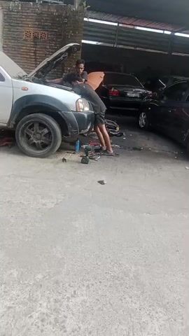 At 00:00, two men are working under the hood of a silver pickup truck in an open-air garage. Tools and car parts are scattered on the ground around the vehicle. In the background, several other cars are parked, including a black sedan, a dark gray hatchback, and another silver pickup truck. A woman with a backpack is standing near the dark gray hatchback at 00:01. By 00:03, the woman is talking to a man standing near the silver pickup truck. The scene appears to be a car repair shop.