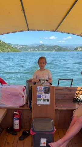 A young girl with pigtails stands at the helm of a wooden boat, holding the steering wheel. She is wearing a white t-shirt that says "TIPS FOR CAPTAIN" and smiles while briefly making a saluting gesture with her right hand. The boat has a yellow canopy overhead. In front of her, the expansive blue water of a lake or river is visible, with distant green mountains under a bright blue sky with scattered clouds. Inside the boat, a red fire extinguisher, two pairs of dark flip-flops, and a grey trash can are on the wooden floor to her left. A white plastic bag filled with items rests on a bench to her immediate left. The lower legs of an adult are visible seated in the lower right corner of the frame. A laminated flyer with text and images is attached to the boat's console below the steering wheel.