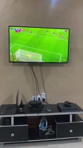 A medium shot shows a TV mounted on a beige wall. A soccer game is displayed on the TV. The score is Chelsea 1, Nottingham Forest 0. Below the TV is a black and white media console. Items on the console include a laptop, a black box, a remote, and a Playstation. In the lower part of the console, there is a blue iron and some cords.