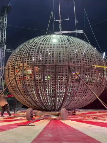 The video shows a "Globe of Death" circus act in progress inside a large, spherical cage. Three motorcycles are visible, each ridden by a person in protective gear, racing around inside the structure. The globe sits atop a wheeled platform, and a spotlight illuminates the performance. The backdrop is a circus tent with red and white stripes on the floor. A person stands outside the cage on the left, seemingly observing the act.