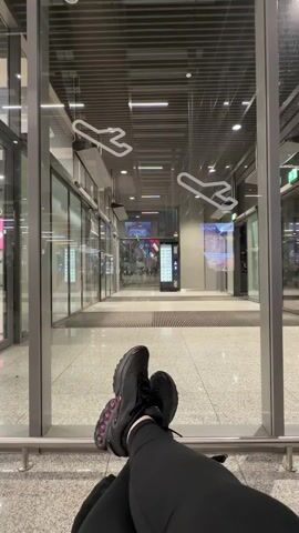 Here's a description of the video:

The video shows a pair of legs in black leggings and black sneakers resting in front of a glass wall. The view through the glass shows an airport hallway with people walking by, some with luggage. The glass has airplane symbols on it. The airport setting is modern with gray flooring and metal accents.