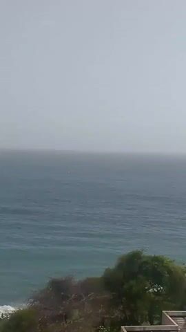 This is a landscape video showing a view of the ocean from a high vantage point. The sky is overcast and hazy. The ocean is a deep blue with some choppy waves. In the foreground, there are some trees and buildings. A small red object, possibly a buoy or a boat, is visible in the water. The video is shaky, indicating it was likely filmed handheld.