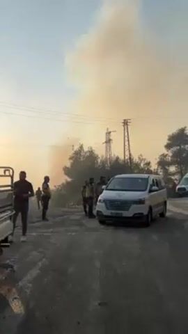 Here's a detailed description of the video:

**Scene:** The video appears to show a roadside scene possibly related to a fire.

**Environment:** The background is dominated by a large plume of smoke rising from what appears to be a fire in the distance. There are trees and power lines in the background as well. The ground appears to be a paved road or rocky surface with vegetation on either side. The sky is partially obscured by smoke, suggesting hazy conditions.

**People:** There are several people visible, mostly standing on the side of the road. Some appear to be wearing what looks like uniform vests or jackets. They are gathered in small groups, possibly discussing the situation or observing the fire.

**Objects:**
*   A white minivan with yellow stripes is parked on the side of the road.
*   A vehicle, likely a pickup truck, is visible on the left side of the frame.
*   Power lines are visible overhead.

**Actions and Interactions:** The people are mostly standing and observing. There is no clear immediate action being taken. The focus appears to be on the distant smoke and the potential fire.

**Overall Impression:** The video seems to capture a scene where authorities or emergency personnel are present at the site of a fire, likely assessing the situation or preparing for action. The presence of uniformed individuals and the minivan suggest a possible response to the fire.