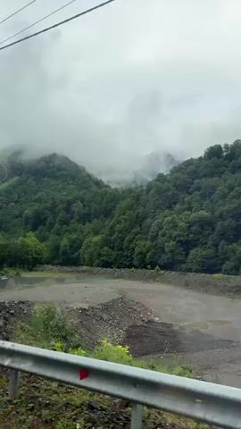 The video shows a view from a moving vehicle. In the foreground, a metal guardrail is visible. Beyond the guardrail is a rocky, barren area with some sparse green vegetation. Further back, a body of water can be seen, reflecting the overcast sky. In the background, lush green forested mountains rise, partially obscured by thick clouds and mist. The scene appears to be a natural landscape, possibly in a mountainous region on a cloudy day. There are no people or distinct structures visible, aside from the guardrail and a glimpse of a corrugated metal fence on the right side of the frame. Power lines are also visible in the upper part of the video. The overall impression is one of a misty, green, mountainous environment.