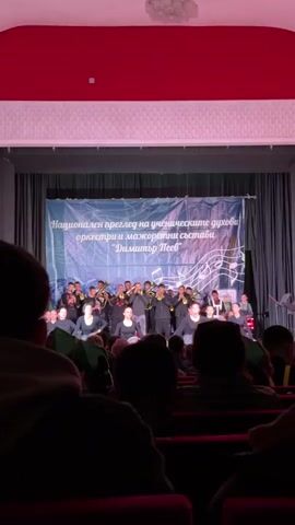 The video shows a performance on a stage in front of an audience. A group of majorettes, wearing short black skirts and black tops, perform with white pom-poms in front of a musical orchestra.  The backdrop banner reads "Национален преглед на ученическите духови оркестри и мажоретни състави 'Димитър Пеев'." which translates to "National review of student brass bands and majorette groups 'Dimitar Peev'."  The audience is seated in red chairs, and only the backs of their heads are visible. The stage is lit, and the venue appears to be a theater or auditorium.