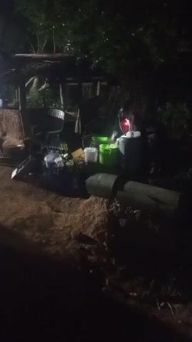The video shows an outdoor gathering at night, likely a small party or cookout. The scene is dimly lit. There's a makeshift structure resembling a bar or serving area made of wood. In front of it, there's a collection of items, including plastic bottles, buckets, and what seems to be a cooler. There are also wooden logs on the ground. People can be seen standing around the bar area, though they are mostly silhouetted. Music is playing in the background. The ground appears to be sandy or dirt.