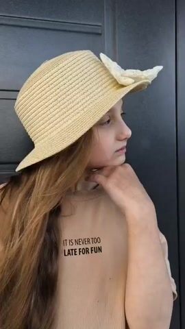 A young girl with long brown hair wearing a straw hat with a white ribbon and a tan t-shirt is standing in front of a dark grey door. She is looking at the camera and her mouth is open. The t-shirt has text on it that says "IT IS NEVER TOO LATE FOR FUN".