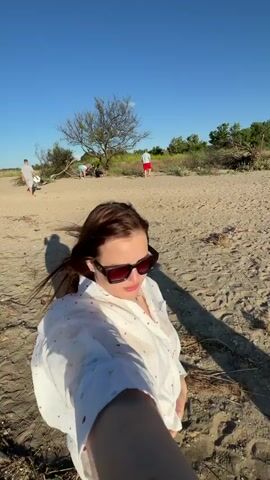 A group of people are at a beach. In the foreground, a woman is taking a selfie. She has dark hair, is wearing sunglasses and a white shirt with red and blue hearts on it. In the background, two people are walking on the beach, and two other people are sitting down near a tree. The scene then shifts to waves lapping on the shore under a clear blue sky. Finally, a person is carrying a baby on their shoulders while walking on the beach towards the sea, with the sun shining brightly.