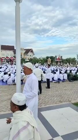 The video captures an outdoor gathering of Muslim worshipers in white attire, seated on prayer mats facing a building with a dome. The scene appears to be in front of a mosque or a similar religious structure. In the foreground, two men in white attire, one with a white skullcap and the other wearing a turban, are visible. One of them is holding a phone, while the other is gesturing with his hand. Behind them, a large crowd of people are assembled, all dressed similarly in white clothing and seated on blue prayer mats. The ground is paved with square tiles. In the background, the building has a prominent dome and intricate architectural details. A tall white flagpole stands near the center of the frame. Several people in dark clothing, possibly security personnel, are also present and appear to be managing the crowd. The sky is overcast.