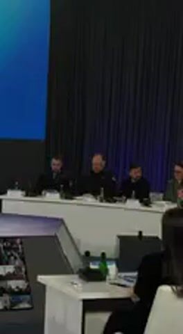 The video captures a formal indoor event, likely an awards ceremony, unfolding in Barvinok, Ukraine. The scene features a long white table with several individuals seated, facing an audience. Microphones, water bottles, and papers are visible on the table. Behind the table, a dark blue curtain serves as a backdrop, illuminated with subtle purple lighting.

In the initial frames, four individuals are seated at the table. One man, wearing a black turtleneck (identifiable as Volodymyr Zelenskyy), stands up and moves towards the center of the stage area, smiling. A woman with glasses and light brown hair, dressed in a green jacket (possibly Olena Zelenska), is seated to his right, also smiling. Another man in a dark suit is seated further down the table.

The foreground shows the back of a person's head, indicating a view from the audience. Visible text on a screen or projected onto the backdrop clearly indicates "Державних нагород" (State Awards) and "Розпочинаємо церемонію вручення Державних нагород" (We are starting the ceremony of presenting State Awards), specifying the nature of the event. The audio reinforces this, with a speaker announcing the beginning of the "ceremony of presenting State Awards." The overall mood appears formal and positive, marked by the smiles of the participants.