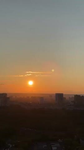 The video captures a time-lapse of a sunset over a city, likely Dikaya Gar', Russia, from a high vantage point. The scene opens with the sun prominent and bright, relatively high in the sky, casting an intense orange glow across the horizon and illuminating scattered clouds. The sky above is a lighter blue-grey.

As the video progresses, the sun gradually descends towards the horizon, its light diminishing but still vibrant. The city skyline is visible below, featuring numerous multi-story buildings and skyscrapers, appearing as dark silhouettes against the glowing sky. A large, dark mass of trees or a forested area is visible in the foreground, further emphasizing the urban landscape.

The colors of the sky evolve throughout the sunset, transitioning from bright oranges and yellows to deeper, more muted hues as the sun dips below the horizon. The distant cityscape and the immediate foreground remain in shadow, enhancing the dramatic effect of the setting sun. No people or specific activities are visible, focusing entirely on the natural phenomenon of the sunset over the urban environment. No text is visible in the video.
