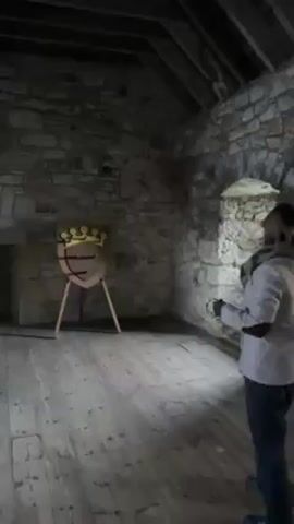 Inside a stone-walled room with a wooden floor and beamed ceiling, a woman throws rope rings at a shield target with a painted crown on it. The target stands on a tripod. The woman wears a grey jacket and dark pants. She throws three rings, one of which lands on the shield. A stone fireplace is visible on the left. A window is on the right.